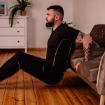 A bearded man in black fitness attire exercising at home, showcasing arm tattoos and a healthy lifestyle.